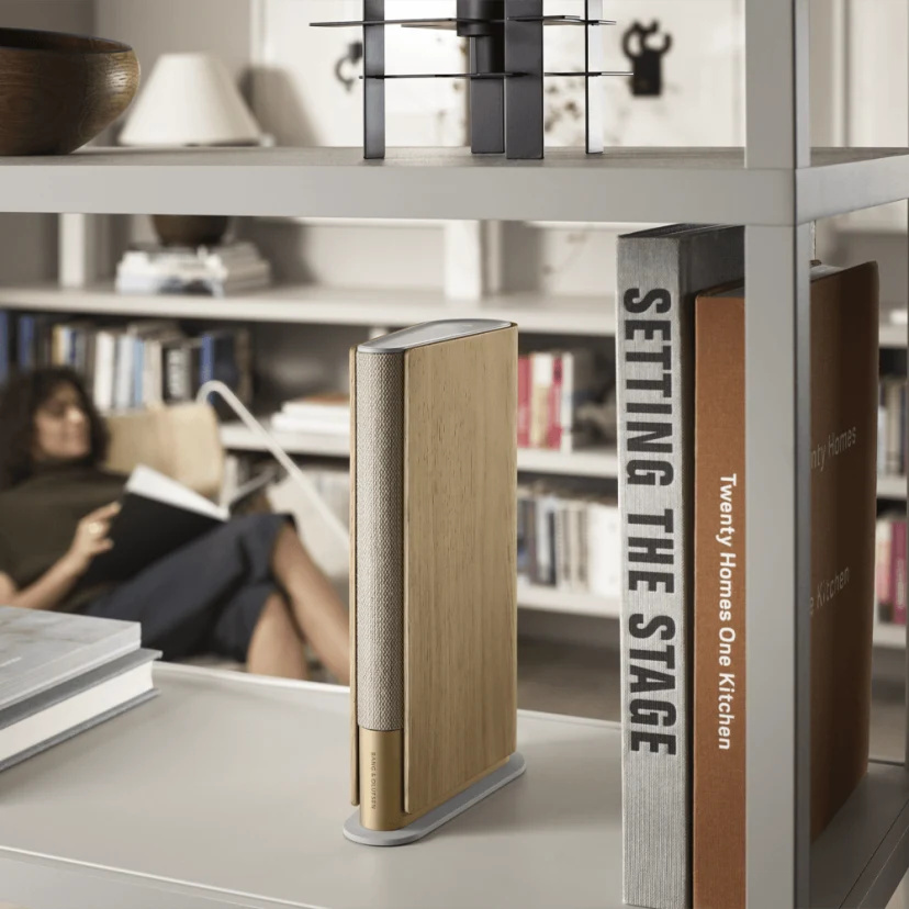 書本外型的碳黑色 Beosound Emerge 揚聲器放在書架上，一名女性在背景一邊聆聽音樂一邊閱讀。